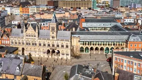 Aerial view of the Guildhall in Northampton. On the left is an older building which has two stories and a clock tower. On the right is a new extension to the building, which also have two floors and glass pain windows. 