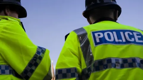 Two police officers are stood with their back towards the camera. They are both wearing a yellow hi-vis jacket. The one on the right has police written on the back.