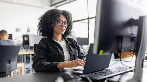Businesswoman wearing black glasses with dark hair, sitting working on computer at her desk in office