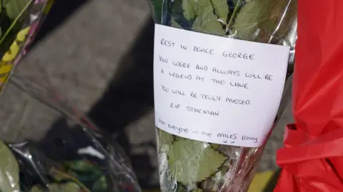 Flowers with a piece of paper which reads: 'Rest in peace George. You were and always will be a legend at the Lane. You will be truly missed. RIP Starman. From everyone in the Miles Family xx'