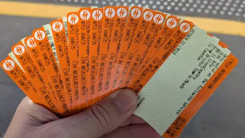 A man standing on a train platform holding a large fan of 15 orange train tickets in his left hand. 