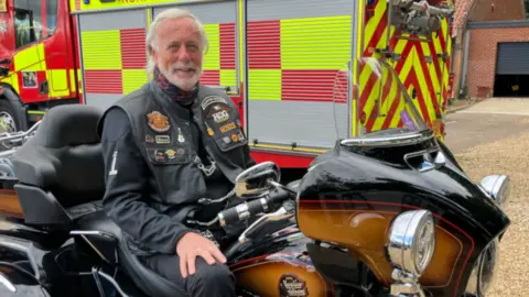 Alex Dunlop/BBC Steve Kimber sat on his motorbike. He is looking directly at the camera and smiling and is dressed head to toe in black. Behind him is a Norfolk Fire and Rescue Service fire engine.