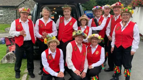 Supplied Eleven members of Furness Morris Dance smiling at the camera back in 2023. They are wearing black trousers, white shirts and red waistcoats, and have hats with flowers at the top.