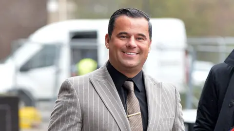 James English wearing a grey and white striped suit, black shirt and brown tie. He is walking into court, smiling at the camera.
