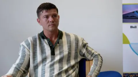 BBC A young man with short dark hair is sitting and looking to the right of the picture. He is wearing a long-sleeved top comprising wide, vertical grey and white stripes. 