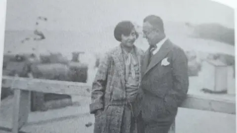 Adrian Kriss An old black and white photograph of a woman with a dark bob, wearing a long coat, standing talking to a man wearing a suit.