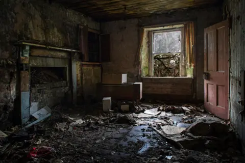 Angus Mackay Photography Plaster and rubbish covers the floor of the room. There is a window with most of the glass missing and red wooden door that is ajar. An old fireplace is partially blocked up.