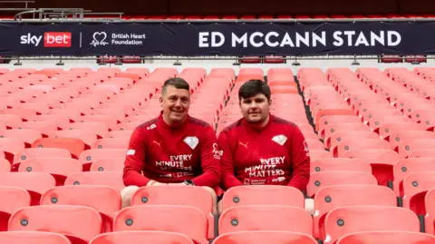 Two men in red shirts sit among red stadium seats. A banner behind them reads Ed McCann Stand