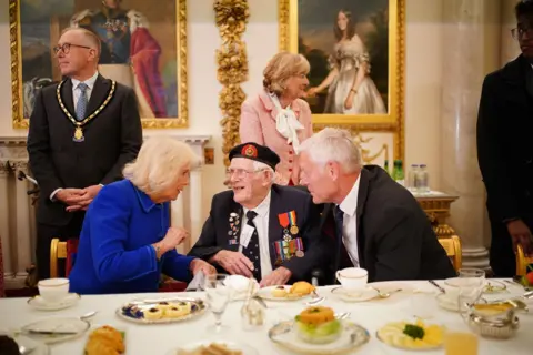 PA Media Queen Camilla speaks with veterans wearing badges and formal attire while they enjoy tea and cakes inside Buckingham Palace. Workers can be seen in the background in front of gilded artwork on the walls.