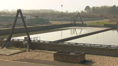 A section of Peel Common wastewater treatment works. There is a short concrete wall in a square, containing brown looking water. In the distance are tankers, a car park and trees. There are triangular structures either end of the water with a metal wire linking them.