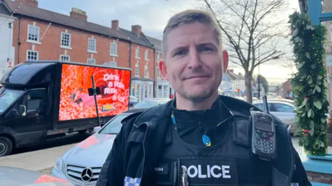 Inspector Robinson looking into the camera, he's wearing police uniform. Behind him you can see Christmas decorations outside a shop in Newport, and over one shoulder there's a van with an anti-drink driving slogan. The slogan reads 'drink a little, risk a lot'.