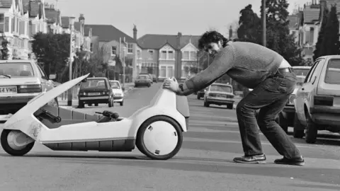 Harry Prosser/Daily Mirror/Mirrorpix/Getty Images A file image of a Sinclair C5 electric tricycle being pushed across a residential street by a man wearing jeans and a pullover. He has short, dark wavy hair. The image is black and white