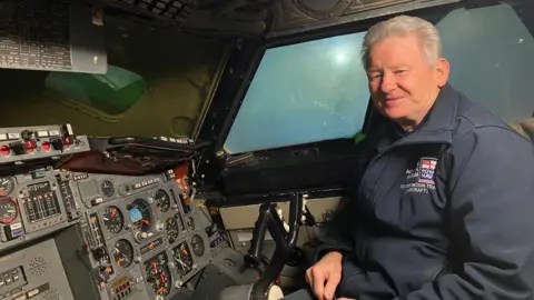 Dave Morris is sitting in the cockpit of the Concorde. He is wearing a blue navy jacket. He's smiling.
