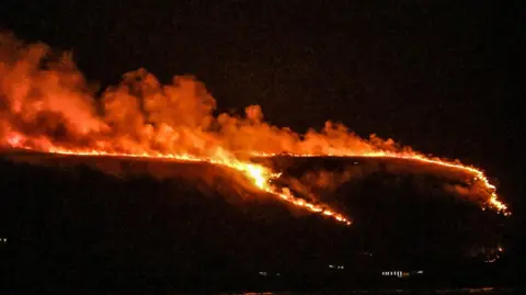 Steve Unwin A line of fire splits into two on a hillside at night. Smoke, tinged orange, billows into the air. Windows in one building at the bottom of the hill are illuminated.