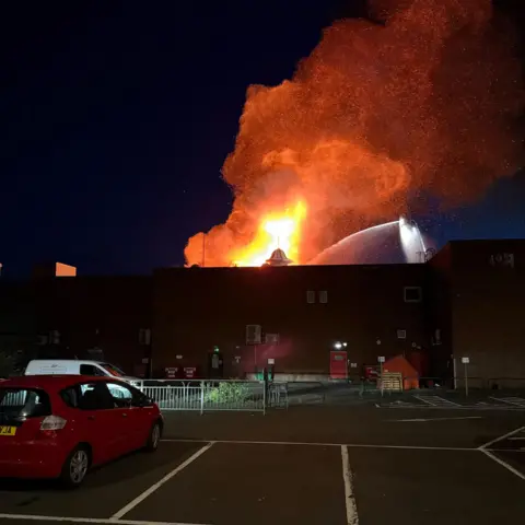 Marc Ferguson The fire from behind a building. A car park is in the foreground. In the upper part of the image, the fire can be seen and a jet of water is being fired at it from above.