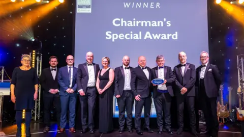 Jack Boskett A line of 10 people dressed in formal evening wear. One of eight men who is wearing a dark suit is holding an award in his hands which is blue and oval shaped with writing on it. 
