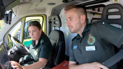 Two paramedics - a man and a woman - sit in the cab of the ambulance taking dispatch instructions