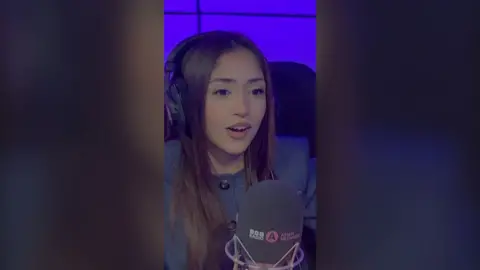 Duaa Karim has long dark brown hair. She is wearing a grey jacked buttoned up to the neck and sitting behind a microphone.
