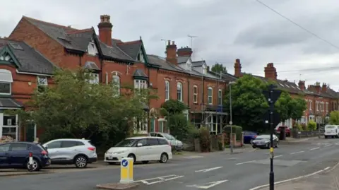 A residential street with red brick houses down one side and cars parked alongside and a road with white and yellow bollard in the middle