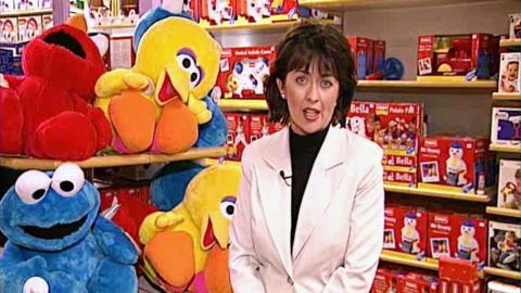 Reporter Denise Mahoney standing in a toyshop, in front of a display of Sesame Street characters.