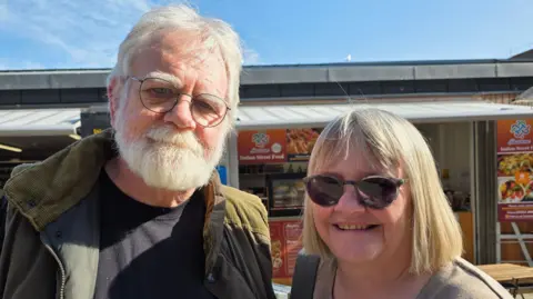 Paul Moseley/BBC David and Ruth Woollard-Kingston are standing in front of a market stall. He is wearing glasses and a green coat over a black t-shirt. She is wearing shades.