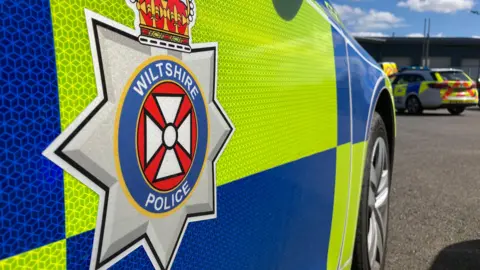 A side-on view of a police car with the Wiltshire Police badge in the foreground.