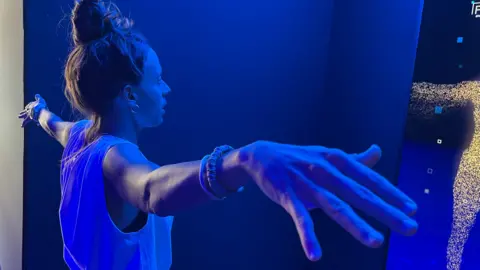 A woman stands with her arms outstretched in front of a digital display