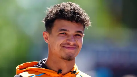 Lando Norris smiles facing the camera. He has curly, short brown hair and the top of his orange hoody can just be seen.