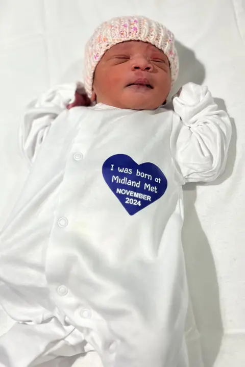 Midland Metropolitan University Hospital A baby wearing a white baby outfit and a knit hat lies on a white sheet. In the words of Babygrow she has a blue heart "I was born in Midland Met in November 2024" written inside.