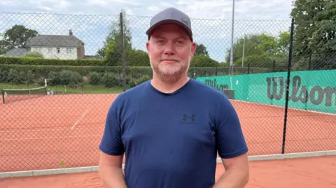 Leanne Rinne/BBC A man standing on a tennis court wearing a blue T-shirt and grey baseball cap. 