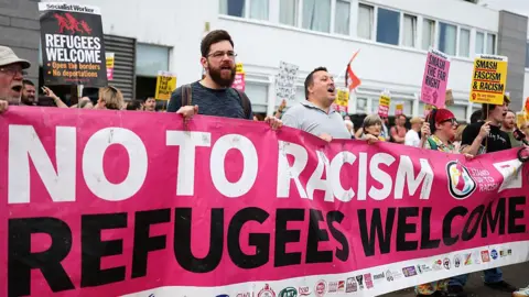 Getty Images A counter-protest outside the Cladhan Hotel in Falkirk on 16 August. A row of people are holding a pink banner which reads No To Racism and Refugees Welcome, and carrying placards.
