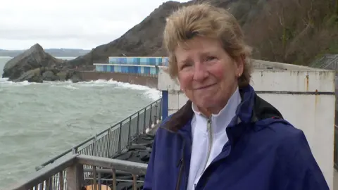Maggie Matthews on the left smiling at the camera. She is wearing a blue coat and a white high neck jumper. 