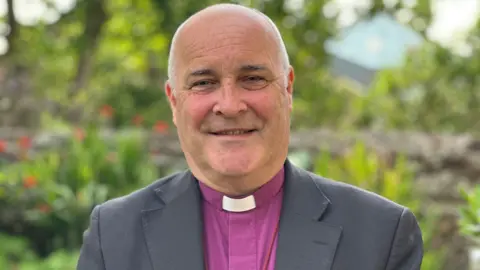 Stephen Cottrell, who has very close cropped grey hair and is wearing a tonsure shirt and dog collar with a grey suit jacket. He is standing in a garden.