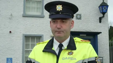 A garda is wearing a yellow and black jacket with a grey strap and a cap. The garda logo is on the jacket and the cap. He's standing in front of a white building with a lantern with the garda logo on it.