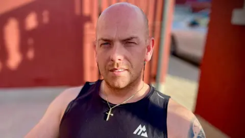 Darren George sits while wearing a gold chain necklace with a cross on it and a black vest top.