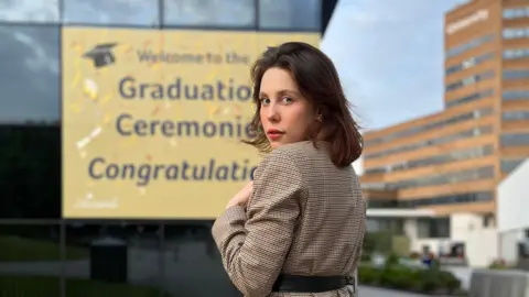 A white female wearing a brown check jacket is at the forefront of the picture looking at the camera. 

There are buildings in the back ground and a sign which is partially covered saying Graduation Ceremonies congratulations. 