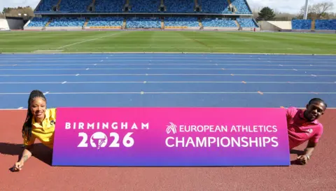 Birmingham 2026 Amber Anning is wearing a yellow T-shirt and has long braided hair she is lying behind a pink and purple sign with the logo of Birmingham 2026 on. At the other end Jeremiah Azu lies behind the sign and is wearing a pink tee-shirt