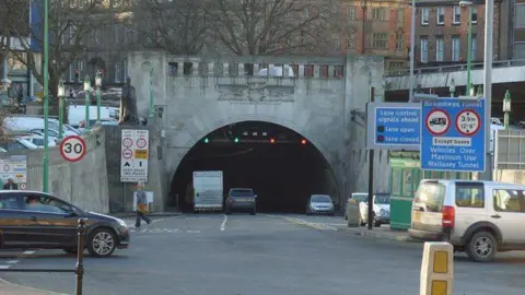 The arched, grey entrance to the Queensway Tunnel, with cars driving into the darkness