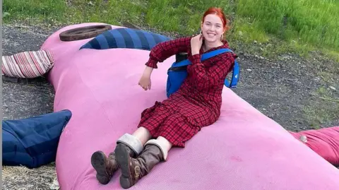 Glasgow School of Art Emilia Evans-Munton, who is wearing a red and black checked dress and brown boots, sitting on a giant pink sock monkey