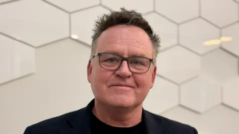 BBC James Tyler, who's wearing a pair of thick-framed glasses and dressed in black, smiles at the camera for a posed head shot. 