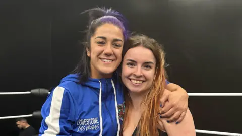 Two woman - Bayley and Natasha Swann - smile for the camera while standing in a wrestling ring. Bayley is wearing a blue tracksuit top saying Lodestone Las Vegas and has dark hair, partly dyed purple. Natasha is shorter, with red hair going past her shoulders. 
