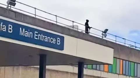 BBC A figure in black can be seen on top of a hospital roof, with railings in front of him. 