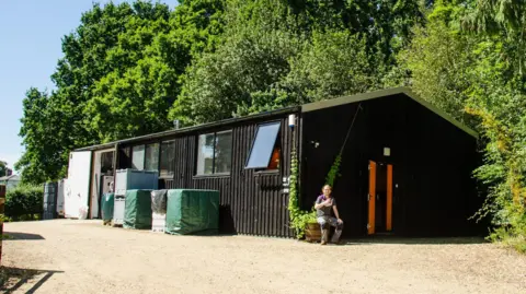 Harry Baynhams A large black barn like structure, with a grey gravel track in front of it, and green shrubbery behind.