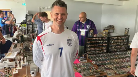 Adam Grocott smiles at the camera, wearing a white Umbro long sleeved England shirt with a red cross on the right shoulder and a blue number 7 in the middle. To his left is a table with hundreds of Corinthians models on, and more to his right. Other tables with memorabilia are behind and people manning the stand or milling about.