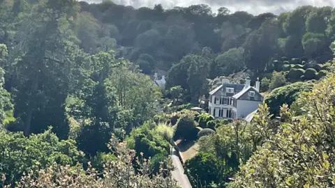 Chateau La Chaire viewed from a nearby hill on a sunny day. It is surrounded by trees. The hotel is large and white with black-rimmed windows.