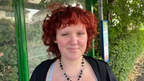 Harriet Copley has red dyed hair with a black cardigan over a blue strapped top with black beaded necklace with a silver necklace attached. Behind her is a green metal bus stop shelter with a timetable board on the right on a lamppost.