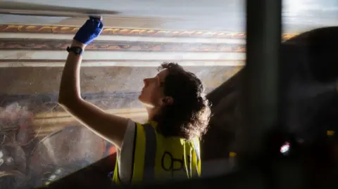 Opus Conservation Woman with high-vis vest on with her left arm stretched up to a ceiling where she is working to restore a painting.