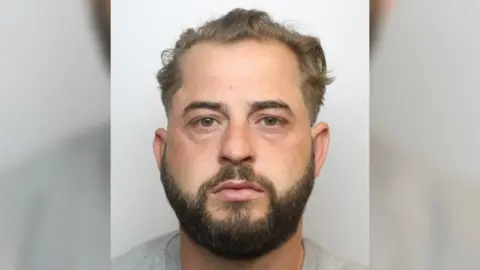 Wiltshire Police A police custody photo of Ashley Sherred. He has brown hair, brown eyes, a dark-brown beard, and is wearing a grey top.