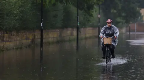 Reuters A female cyclist pedals though flood water on a road. The woman wears a clear plastic rain poncho, black clothes and a pink helmet. Her bicycle has a wicker basket. Water has filled the road and surrounded lampposts. 