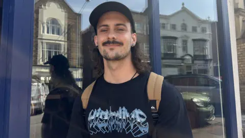 BBC/Chloe Hughes A man with long brown curly hair under a black cap, and a moustache. He is wearing a black top with a brown rucksack on his back. He is smiling.